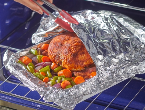 Person using tongs to pull back aluminum foil to view a whole roasted chicken in an aluminum foil lined roasting pan