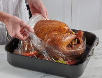 Person removing a turkey oven bag from a fully roasted turkey sitting in a roasting pan