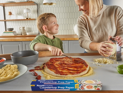 Mother and son making a pizza with Reynolds Kitchens Countertop Prep paper laid out on the counter to avoid a mess. Sauce and toppings are scattered everywhere but the paper is keeping the counter clean.