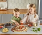 two kids topping a homemade pizza that is sitting on a counter topped with countertop prep paper
