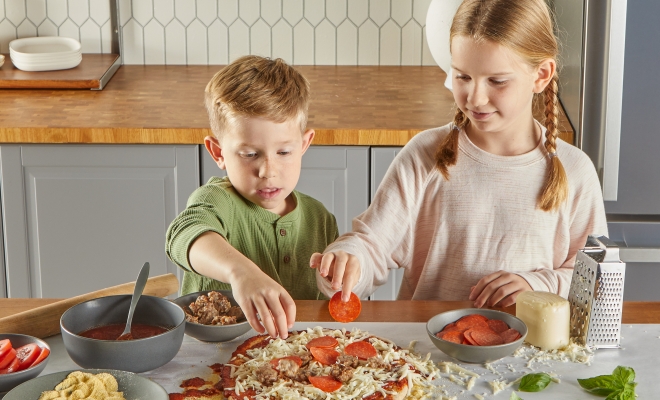 two kids topping a homemade pizza that is sitting on a counter topped with countertop prep paper