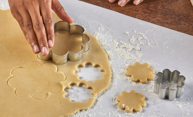 person using a cookie cutter to shape cookie dough that is rolled and sitting on a sheet of countertop prep paper