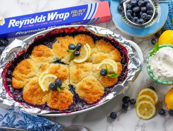 Lemon Blueberry Cobbler in an aluminum foil lined casserole dish sitting next to a box of Reynolds Heavy Duty Foil, fresh lemons and blueberries